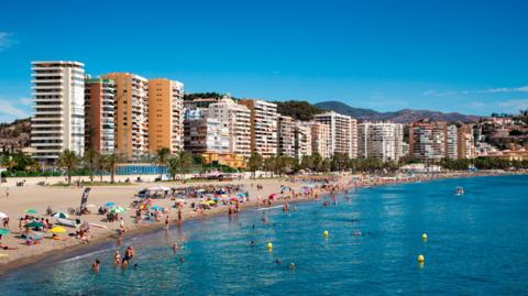 A sunny beach scene with people sunbathing on the shore and some people swimming. There are high-rise buildings in the background.