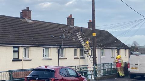 Firefighters are the scene of the suspected gas explosion. One firefighter is up a ladder outside a row of terraced houses, while two others look on. Several roof tiles appear to be missing from the roof of a mid-terraced house. 