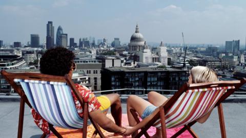 Two people holding hands whilst seated in brightly coloured deckchairs on a rooftop looking towards the London skyline with St Paul's cathedral in the distance 
