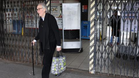 A man carrying a shopping bag out of a Tube station which has the shutters partly closed