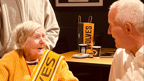 A woman with white hair wears an orange jumper, which is the same colour as the Wolves kit, with a sash that says Wolves. She is looking at and chatting to Steve Bull, who has white hair and is wearing a white shirt. Behind them is a table with a bag and some paper cups that have the Wolves logo on them. 