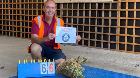 Curtis Leach holding up a sign that says "new world record" as he kneels down next to a very big - record - celeriac and a sign that says "6.6". 