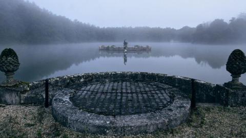 A mist-covered lake at Witley Park is viewed from a stone terrace. At the centre of the water is a platform topped by a statue of Neptune, marking the location of the underwater room hidden beneath the surface.