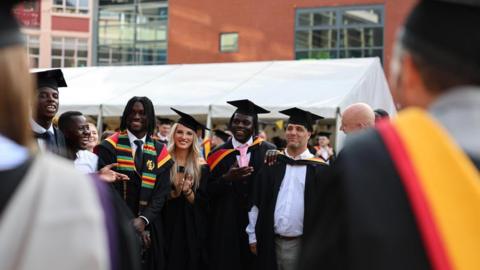 A group of graduates stand in black gowns and caps, smiling at each other. They have red and yellow hoods on the top of their gowns. There are six men and one woman in the group in the middle of the photo. A white tent is behind them.