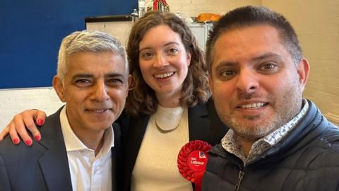 A woman wearing a red Labour rosette stands in between two men. She has curly hair and is smiling. Sadiq Khan, on the left, has grey hair, and wears a white open necked shirt and grey blazer. The man on the right wears a floral shirt and black puffer jacket.