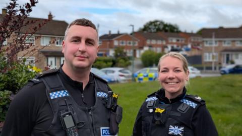 A male and a female police officer stand side-by-side in black uniform. They are stood on a housing estate and with properties and grass behind them. They are smiling at the camera. 