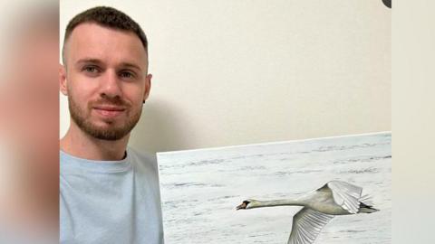 Michael Velychko, who has short, brown hair and a beard and is wearing a grey sweatshirt, is holding a painting of a swan gliding across water. 