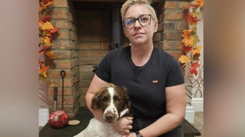 A woman with short blonde hair and glasses is kneeling in front of a fire place. She is wearing a black T-shirt and black bottoms and is cuddling a white springer dog with brown markings.