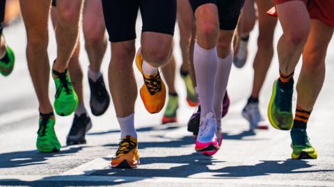 A close-up of a group of runners' legs. They are wearing bright-coloured running trainers and are running on a road.