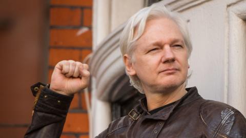 Julian Assange in brown leather jacket with right fist raised