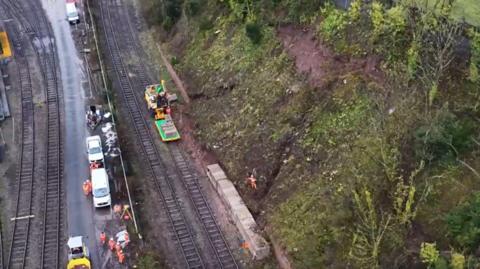 An overhead image of workers on a railway line who are repairing a damaged embankment on the right. Concrete blocks have been installed at the bottom of the embankment.