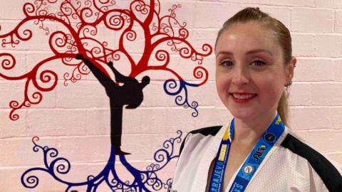 A woman wearing a white jacket with black stripes on the shoulders stands in front of a wall. She is wearing a blue lanyard. Behind her is the painting of a red and blue tree, featuring the silhouette of a man kicking his leg into the air.