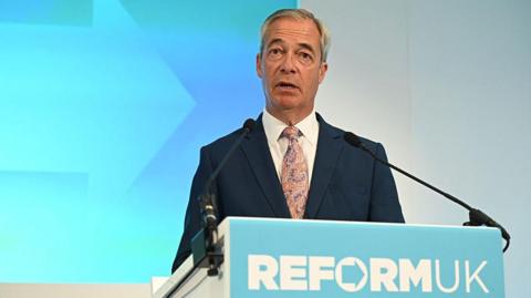 Nigel Farage standing a podium. He is wearing a navy suit and purple and beige tie.  He is standing in front of a light blue and white wall. The podium in front of him reads Reform UK, in the form of the party's logo.