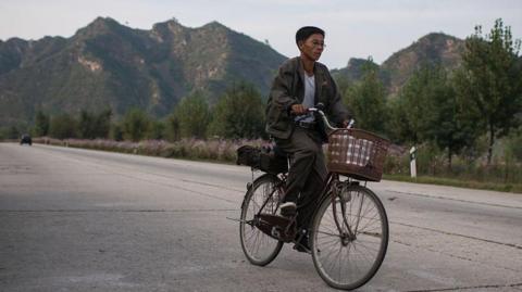 A man in a sweater rides a bicycle down a highway in front of a mountainous background