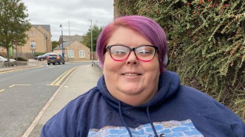 A close up of Liz Owen. She has short purple hair, glasses with a pink and black rim. She wears a navy hooded top. Behind her is a green bush and a road with double yellow lines.