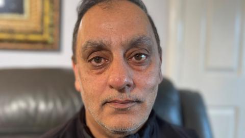 Araf Saddiq stares at the camera with a serious, sad expression, He is wearing a black jacket zipped up to the neck. He is sitting on a leather couch in a domestic setting with a gold framed painting visible in the background.
