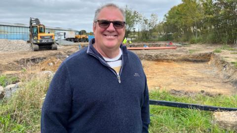 A smiling man in a blue fleece and sunglasses stands in front of a building site where grass has been removed. A digger is in the background.