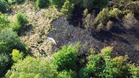 An aerial image of a country park showing grassland and trees surrounding it. Park of the grassland has been scorched black by a fire. Firefighters can be seen working with one spraying a hose on the ground.