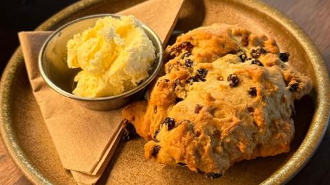 A crumbly baked good with dried fruit on a plate next to small bowl of clotted cream.