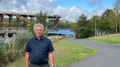 Councillor Gary Haley on the Riverside Row path. He has grey hair and is wearing a navy short-sleeved shirt. Grass and trees are either side of the path, while Dunston Staiths - a wooden structure once used to load coal on to ships on the river - can be seen in the background.