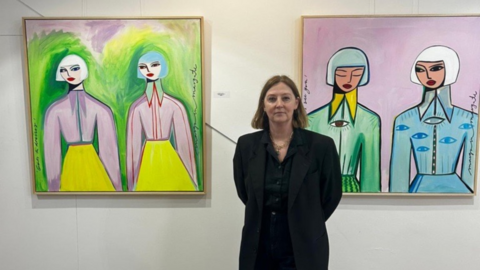 Margo McDaid dressed in black, stood in front of a wall with two of her paintings. The works each depict two women and are in pink, green and blue colours.