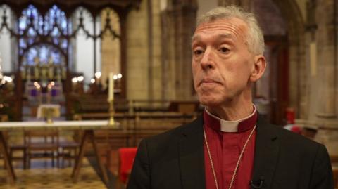 Andrew John in a red shirt and black blazer with a dog collar, looking at the camera in front of an altar. He has grey hair and brown eyes. 