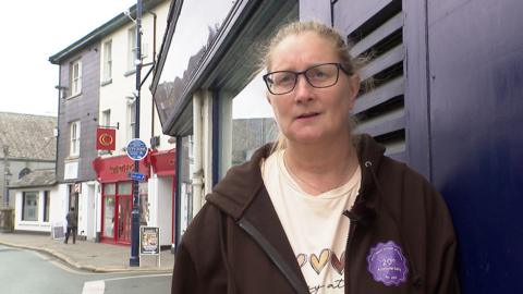 Chrissie Read has her hair tied back and is standing outside on a pavement leaning against the wall of a shop, wearing glasses, a cream t-shirt and a black hoodie. 