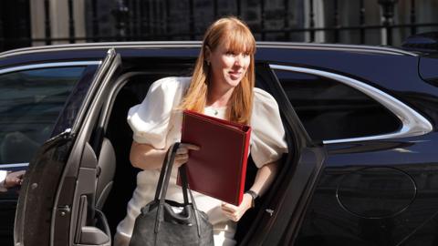 Angela Rayner steps out of a black car wearing a white dress. She carries a red folder and a black bag.