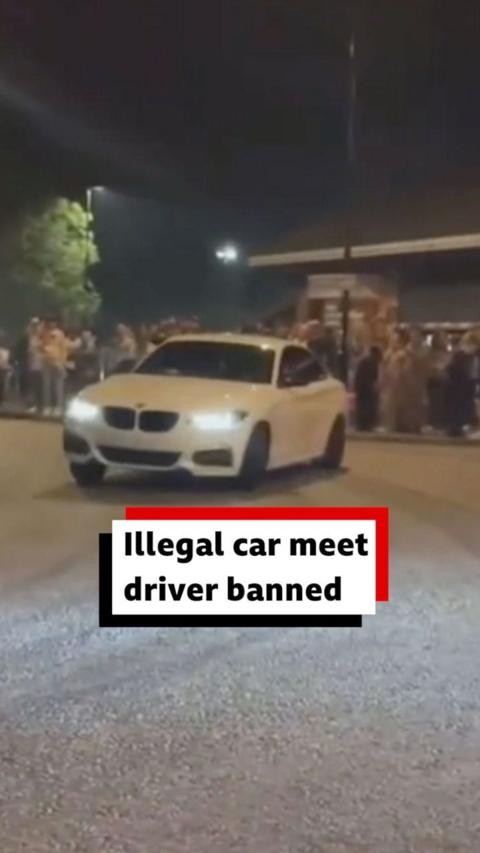 A white BMW drives along the road at night, there are crowds of people in the background