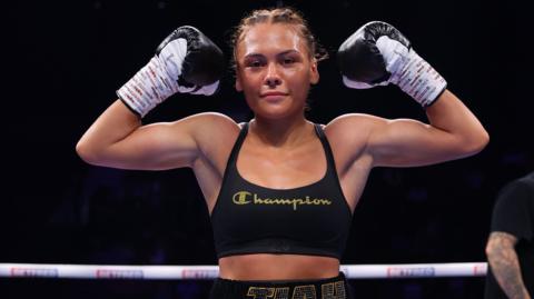 Tiah -Mai Ayton flexing her biceps while looking into the camera. She is standing in the boxing ring wearing a black sports bra, black boxing gloves with her hair plaited into tight cornrows. 