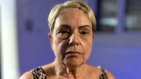 A head and shoulders picture of Linda Stokes who is looking at the camera with a straight face. Behind her the background is blurry. It appears to be a white wall with blue light shining on it and a window.