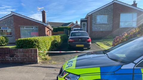 Detached bungalow with two cars in the driveway and police tape across the drive. A police car sits outside the house.