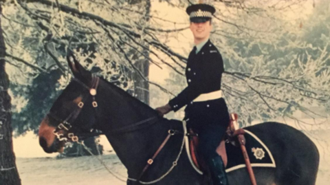 A man in police uniform is sat on the back of a dark brown horse which has a police badge on its saddle rug. The man's head is turned towards the camera while the horse is stood in profile.