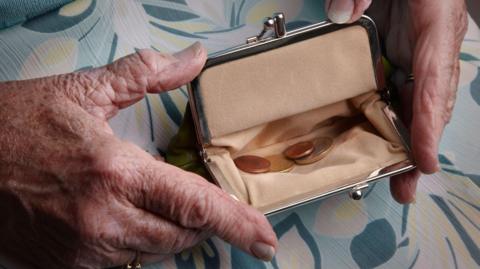 An image of an older woman's hands holding a small purse - it is open and a handful of coins can be seen inside it. The hands and purse are resting on the woman's lap and she is wearing a teal dress or skirt with a floral pattern.