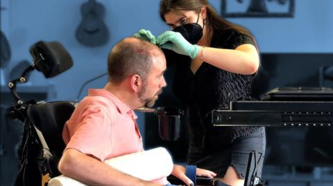 Lady wearing face mask attaches something to the head of a man using a wheelchair