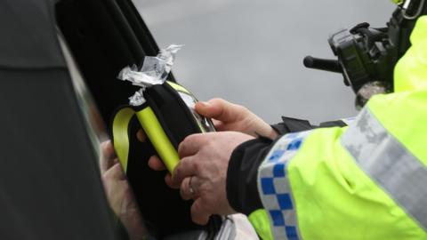 A close up of a police officer in a luminous yellow jacket stood at a car window. The officer is holding a breathalyser test and console. The car window is wound down.