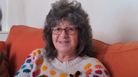 Kitty wearing a jumper with rainbow coloured polka dots. She has mid-length dark hair and is wearing glasses. She is sitting on an orange sofa.