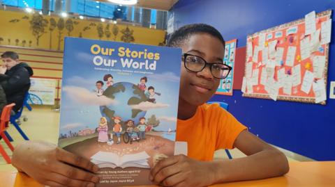 A young boy in an orange t-shirt is sitting in a classroom holding a book which reads, Our Stories, Our World