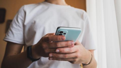 An unidentifiable young man wearing a white t-shirt is shown holding his smartphone in his hands.