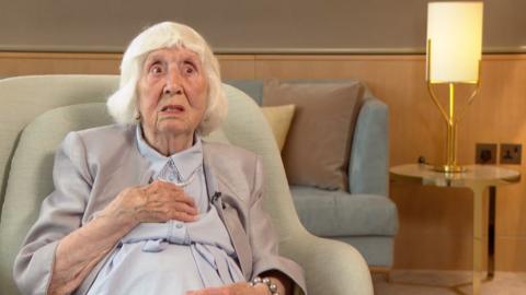 Betty Brown wearing a blue dress, grey jacket and pearls sits in a green armchair. In the background is a lamp on a table.