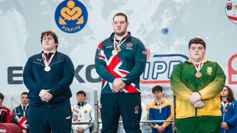 Three young men on a podium, with two of them wearing blue tracksuits and the third a yellow and green tracksuit, with medals around their necks