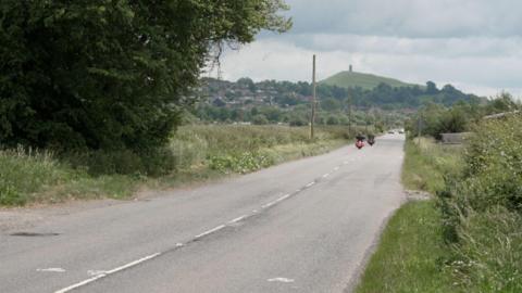 A straight country road with motorcyclists driving along and cars further away. Beyond the trees in the far distance is Glastonbury Tor.