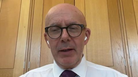 A bald man with black glasses and white shirt and claret tie sat in an oak panelled room