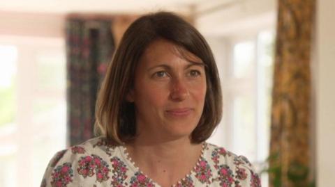 Kate, a woman with short brown hair and wearing a pink floral dress. She is smiling. The background is out of focus but a green house plant and some curtains can be seen. There are also white window frames and it looks to be a bright day outside.