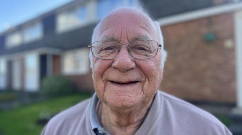 A head and shoulders shot of a man with grey hair and glasses. He is smiling at the camera and wearing a lilac jumper with a blue shirt collar poking out. He is standing in front of a row of houses. 