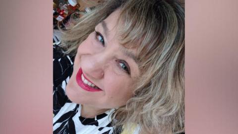 A close-up selfie of a woman with blonde hair and bright pink lipstick smiling at the camera. The woman is wearing a black and white top. She appears to be standing in a kitchen and there is a bottle of ketchup behind her.