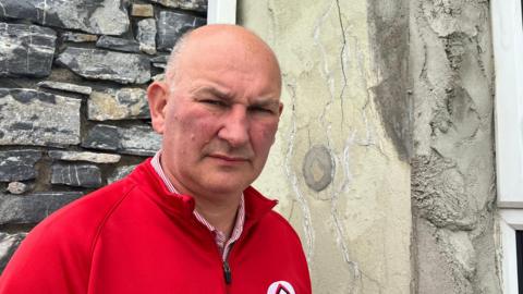 Ali Farren is wearing a red fleece and a white and red striped shirt.
He is standing near a house with defective concrete blocks.