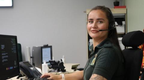 Nicole Douglas is sat at a desk looking at the camera. She has long, dark hair and is wearing a headset and a green shirt. There is a telephone on the desk as well as various monitors.