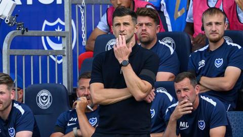 John Mousinho stands in front of the home dugout with his left hand in front of his mouth