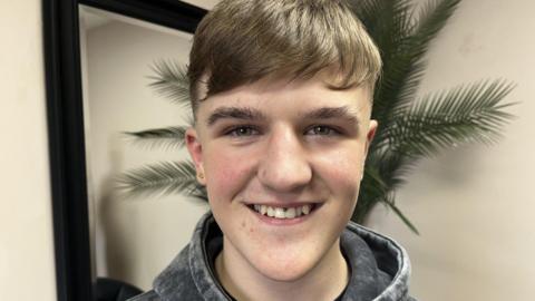 A teenage boy with brown hair is smiling at the camera,
He has a number of facial piercings